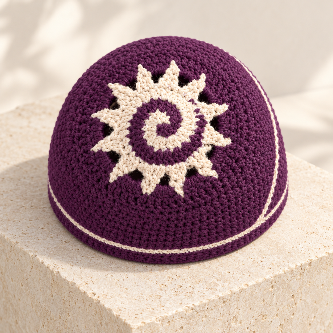 Plum crochet beanie with an ivory spiral sun motif, pointed rays, and a fine ivory edge stripe, displayed on a pale stone plinth.