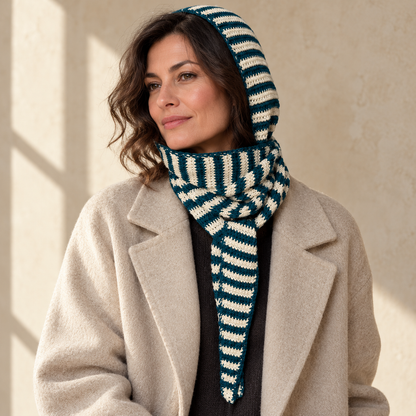 Adult woman wearing a teal-and-ivory striped crochet hood scarf with a wrapped neck and pointed end over an oatmeal coat against a beige studio wall.